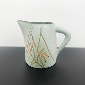 Può includere: Un'anfora in ceramica verde chiaro con un disegno marrone di piante di bambù stilizzate. L'anfora ha un corpo arrotondato e un manico.