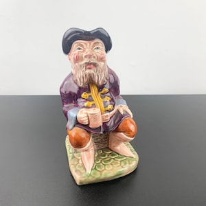 Puede incluir: Una figurita de cerámica de un hombre que lleva una chaqueta morada y dorada, pantalones marrones y un sombrero negro. Está sosteniendo una taza en su mano derecha y sentado en una base verde y marrón.