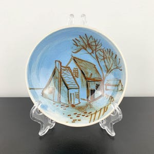 May include: A small, round ceramic plate with a hand-painted landscape scene. The plate features a light blue sky and a brown and white illustration of a house and tree. The plate is displayed on a clear plastic stand.
