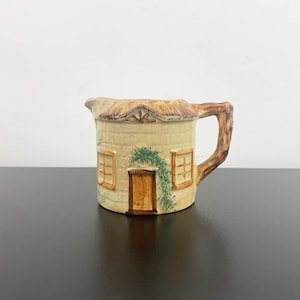 May include: A ceramic creamer in the shape of a cottage with a thatched roof, brown door, and two windows. The creamer is painted in shades of brown, beige, and green.