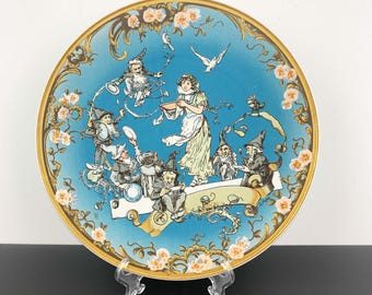 May include: A blue and gold ceramic plate with a whimsical illustration of a girl and seven dwarves. The plate features a blue background with gold accents and floral details. The illustration depicts the girl holding a bowl and the dwarves holding various objects, including a drum, a spoon, and a cup.