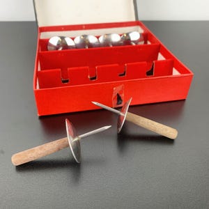 May include: A set of vintage silver-toned upholstery tacks with wooden handles, displayed in a red box. The box contains several additional tacks with rounded silver tops. The tacks are designed for securing fabric to furniture.
