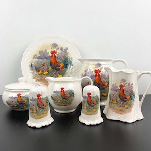 Puede incluir: Un juego de vajilla de cocina de cerámica blanca con un diseño de gallo y pollitos. El juego incluye una jarra grande, una jarra pequeña, un azucarero, un plato y dos saleros y pimenteros.