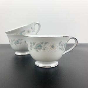 May include: Three white teacups with a floral design in shades of green and white. The cups have a silver rim and a delicate floral pattern.