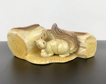 May include: A ceramic planter shaped like a log with a squirrel sitting on top. The planter is yellow with brown accents and has two separate planting areas.