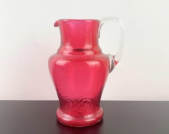 May include: A red glass pitcher with a clear glass handle. The pitcher has a textured surface and a rounded base.