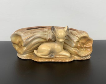 Może przedstawiać: Ceramiczna doniczka w kształcie pnia drzewa z figurką jelenia siedzącego na górze. Doniczka jest brązowa i beżowa z szkliwioną powierzchnią.