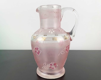May include: A pink glass pitcher with a clear handle and spout. The pitcher is decorated with white and gold bands, and pink floral designs. The pitcher is likely used for serving beverages.