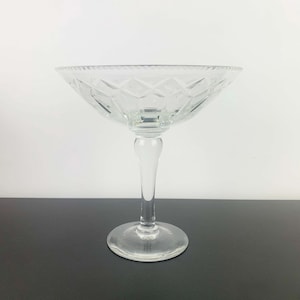 Puede incluir: Un cuenco de postre de cristal transparente con pie. El cuenco tiene un diseño ancho y poco profundo con un patrón de diamantes y un borde festoneado. El tallo es delgado y elegante, y la base es redonda. El cristal es transparente, permitiendo el paso de la luz.