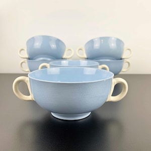 May include: A set of six light blue soup bowls with cream-colored handles. The bowls have a crackle glaze finish and are arranged on a dark surface against a white background. The bowls are stacked in pairs.