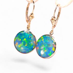 May include: A pair of gold-colored leverback earrings, each featuring a round, iridescent opal stone. The opals display a vibrant mix of blue, green, and yellow hues, set in a polished gold frame. The earrings are suspended against a white background.
