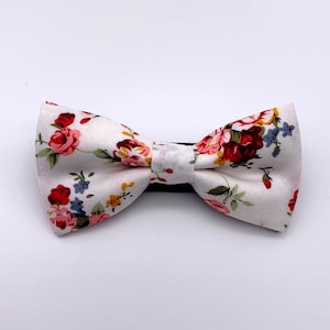 Könnte beinhalten: Eine weiße Fliege mit einem Blumenmuster aus roten, rosa und blauen Blumen. Die Fliege hat ein schwarzes Band und ist vor einem weißen Hintergrund platziert. Accessoire für formelle Anlässe.