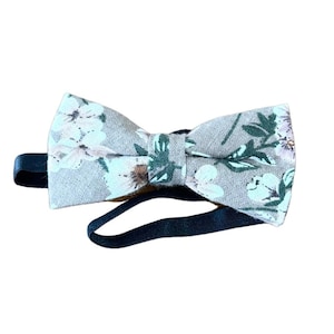 May include: A light brown bow tie with a floral pattern in shades of green, white, and pink. The bow tie has a black adjustable strap.