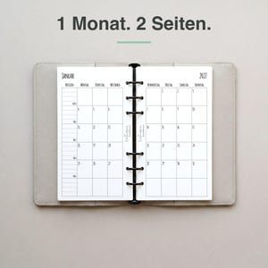 Könnte beinhalten: Ein offener Planer mit hellgrauem Einband zeigt einen Monatskalender für Januar 2027 auf zwei Seiten. Der Text 1 Monat. 2 Seiten. steht oben. Der Planer hat in der Mitte eine schwarze Ringbuchmechanik mit sechs Ringen aus Metall.