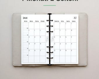 May include: An open planner with a light gray cover featuring a monthly calendar layout for January 2027 across two pages. The text 1 Monat. 2 Seiten. appears at the top. The planner has a black six-ring metal binder mechanism in the center.