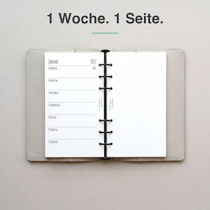 Könnte beinhalten: Ein offener Ringbuch-Planer mit hellgrauem Einband. Die linke Seite zeigt einen Wochenplan für Januar 2027 mit deutschem Text, die rechte Seite ist punktkariert. Über dem Planer steht der Text 1 Woche. 1 Seite.