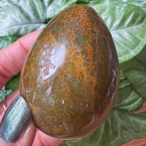 Puede incluir: Un huevo de ágata verde y marrón con una superficie lisa y pulida. El huevo tiene un patrón único de vetas marrones y blancas.