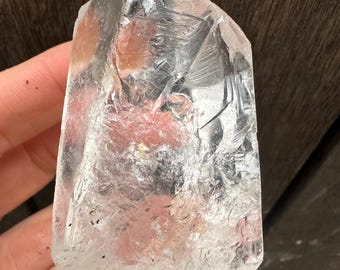 Puede incluir: Un cristal de cuarzo transparente con una textura natural rugosa. El cristal se sostiene en una mano, mostrando su cualidad translúcida y la luz que lo atraviesa. La forma del cristal es irregular, con una parte superior puntiaguda y una base dentada.