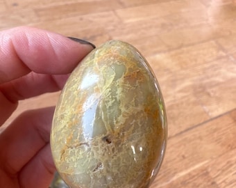 May include: A polished, egg-shaped stone with a mix of green and tan hues. The stone has a glossy finish and visible natural patterns. The egg is held in a hand, suggesting its size and shape. The stone is likely a decorative or collectible item.