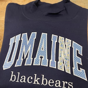 Könnte beinhalten: Ein marineblaues Sweatshirt mit dem Schriftzug UMAINE in großen, hellblau gemusterten Stoffbuchstaben mit weißem Rand und dem Wort blackbears, das in weißen Kleinbuchstaben unter dem Haupttext eingestickt ist.