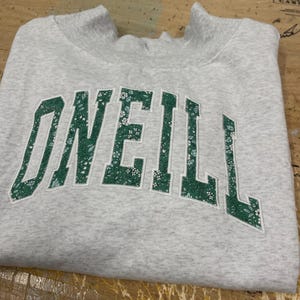 May include: A light gray heathered crewneck sweatshirt featuring the word O'NEILL in large, arched, block-style letters. The lettering is filled with a green and white floral pattern and outlined in white embroidery thread.
