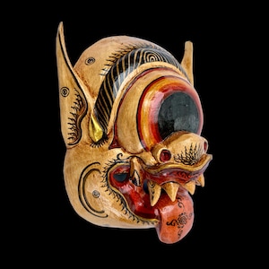 May include: A detailed, hand-carved wooden mask with a fierce expression. The mask features a tan base with red, black, and gold accents. The eyes are large and round, with red and black details. The mouth is open, revealing sharp teeth and a red tongue. The mask has large ears and is set against a black background.