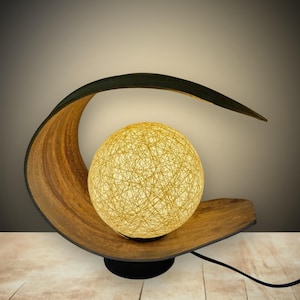 May include: A table lamp featuring a curved, dark brown wooden base that arches over a spherical light shade. The shade is constructed from a dense, intricate web of thin, light yellow fibers that emit a warm, soft glow when illuminated.