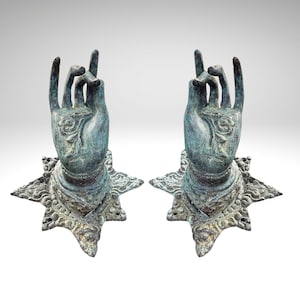 May include: Two bronze-effect hand sculptures in a mudra pose, with detailed floral patterns on the palms and star-shaped bases. The hands are raised, with the index and middle fingers extended. The sculptures have a weathered, antique finish.
