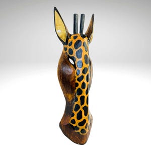 May include: Wooden giraffe mask with black and orange spots. The mask is carved from wood and painted with a realistic giraffe pattern.