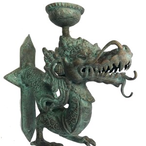 May include: A bronze-colored dragon-shaped wall sconce with a cup-shaped holder. The dragon has intricate details, including scales, teeth, and curled appendages. The sconce has a green patina, suggesting age or exposure to the elements.