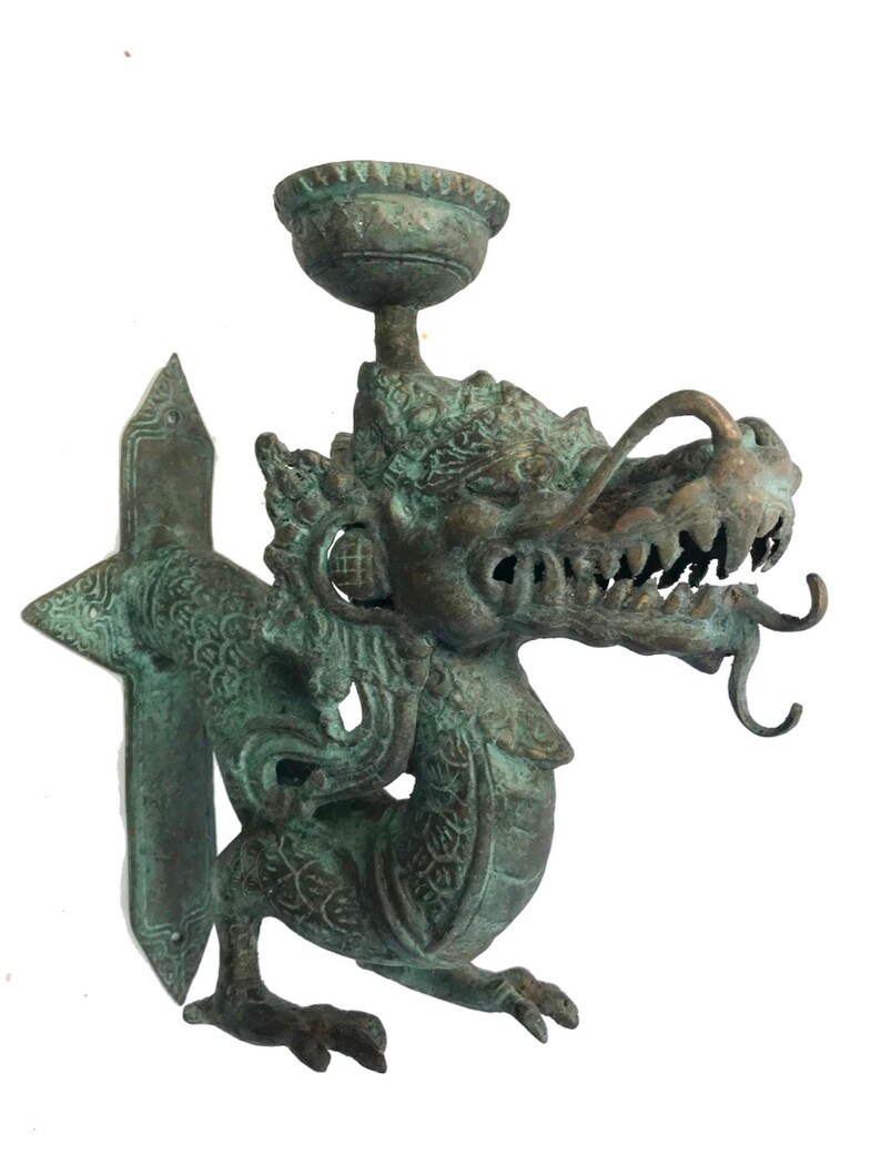 May include: A bronze-colored dragon-shaped wall sconce with a cup-shaped holder. The dragon has intricate details, including scales, teeth, and curled appendages. The sconce has a green patina, suggesting age or exposure to the elements.