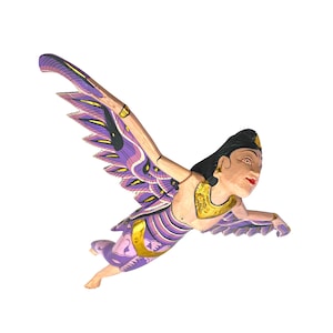 May include: A wooden sculpture of a winged figure in flight. The figure has a painted face, black hair, and wears a gold-colored necklace. The wings are purple and gold, with intricate feather details. The figure's clothing is also purple and gold.