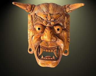 May include: A detailed, wooden mask with a fierce expression. The mask features prominent horns, a wide open mouth with visible teeth, and intricate carvings. The color is a warm, natural wood tone, suggesting a handcrafted item. The mask is likely for decorative or ceremonial purposes.