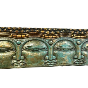 May include: A rectangular, teal-colored wooden wall hanging featuring three carved Buddha faces. The piece has gold and black accents along the top and around the faces, creating a striking contrast. The faces have closed eyes and serene expressions.