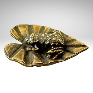May include: A bronze frog sculpture sitting on a leaf. The frog is detailed with textured skin and has a realistic pose.
