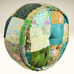 May include: A round patchwork cushion with a button in the center. The cushion is made of various fabrics in shades of green, blue, yellow, and brown. The fabrics have floral and geometric patterns.