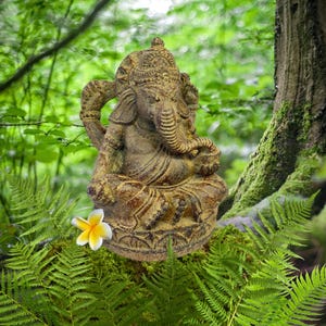 May include: A golden brown Ganesha statue sitting on a lotus base, surrounded by green ferns and a white flower. The statue is intricately detailed with a crown and a trunk.