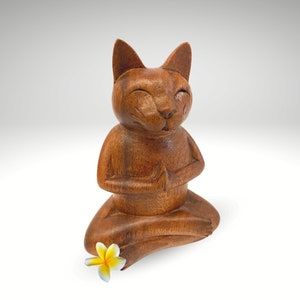 Op de afbeelding: Een houten kattenbeeldje dat in meditatieve houding zit met zijn handen in elkaar. De kat heeft een bruine houten afwerking en een kleine gele bloem aan zijn voeten.