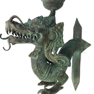 May include: A bronze-colored dragon sculpture with a candle holder. The dragon has detailed scales, sharp teeth, and a long, curling tongue. The sculpture has a weathered, aged appearance, with hints of green patina. The candle holder is a small, round cup above the dragon's head.