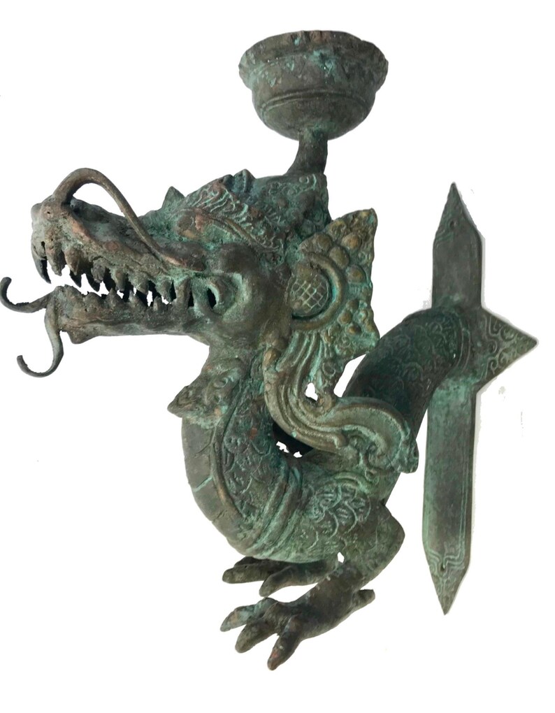 May include: A bronze-colored dragon sculpture with a candle holder. The dragon has detailed scales, sharp teeth, and a long, curling tongue. The sculpture has a weathered, aged appearance, with hints of green patina. The candle holder is a small, round cup above the dragon's head.