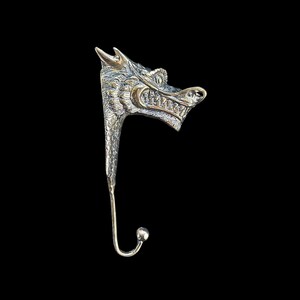 Puede incluir: Un gancho de metal plateado con forma de cabeza de dragón. El dragón tiene escamas detalladas, una boca abierta con dientes y un gancho curvo en la parte inferior. El fondo es negro.
