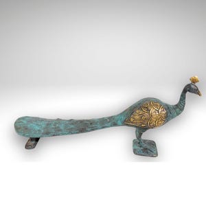 May include: A metal peacock figurine featuring a weathered teal and green patina finish across its long tail and body, contrasted by polished gold-tone metal accents on the wing and head crest, standing on a square base with a tail support.
