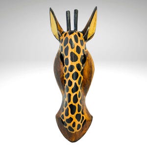May include: A wooden giraffe head wall decor with a brown and black painted pattern. The giraffe head is carved from wood and has a realistic appearance.