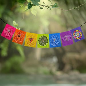 May include: A string of seven rectangular fabric flags hanging horizontally. Each flag features a distinct color and a unique chakra symbol in white or multi-colored ink, including pink, orange, yellow, green, blue, purple, and violet.