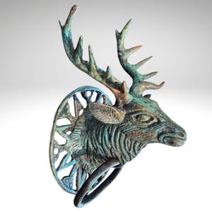 May include: A bronze-colored deer head door knocker with a weathered, verdigris finish. The knocker features detailed antlers, a circular backplate with an openwork design, and a ring for knocking. The overall design is rustic and decorative.