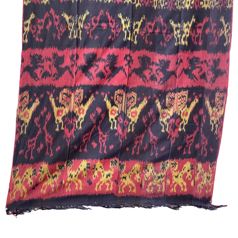May include: A black, red, and gold woven fabric with a repeating pattern of stylized animals. The fabric has a fringed edge.