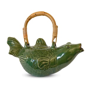 May include: A green ceramic teapot shaped like a fish, featuring a woven rattan handle. The teapot has a detailed scale pattern and a small lid. The fish's mouth is open, and the tail fin is visible.