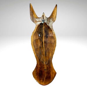 May include: A wooden mask with a carved face and a pointed top. The mask is painted in shades of brown and has a distressed look. It has a metal hook on the back for hanging.