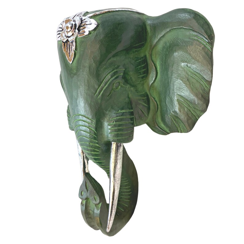 May include: Green wooden elephant head wall decor with silver accents. The elephant has a detailed carved design and a floral pattern on its forehead.
