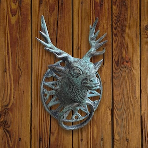 May include: A bronze deer head door knocker with antlers and a circular backplate. The knocker is mounted on a wooden door with vertical planks. The deer head has detailed features and a ring for knocking.